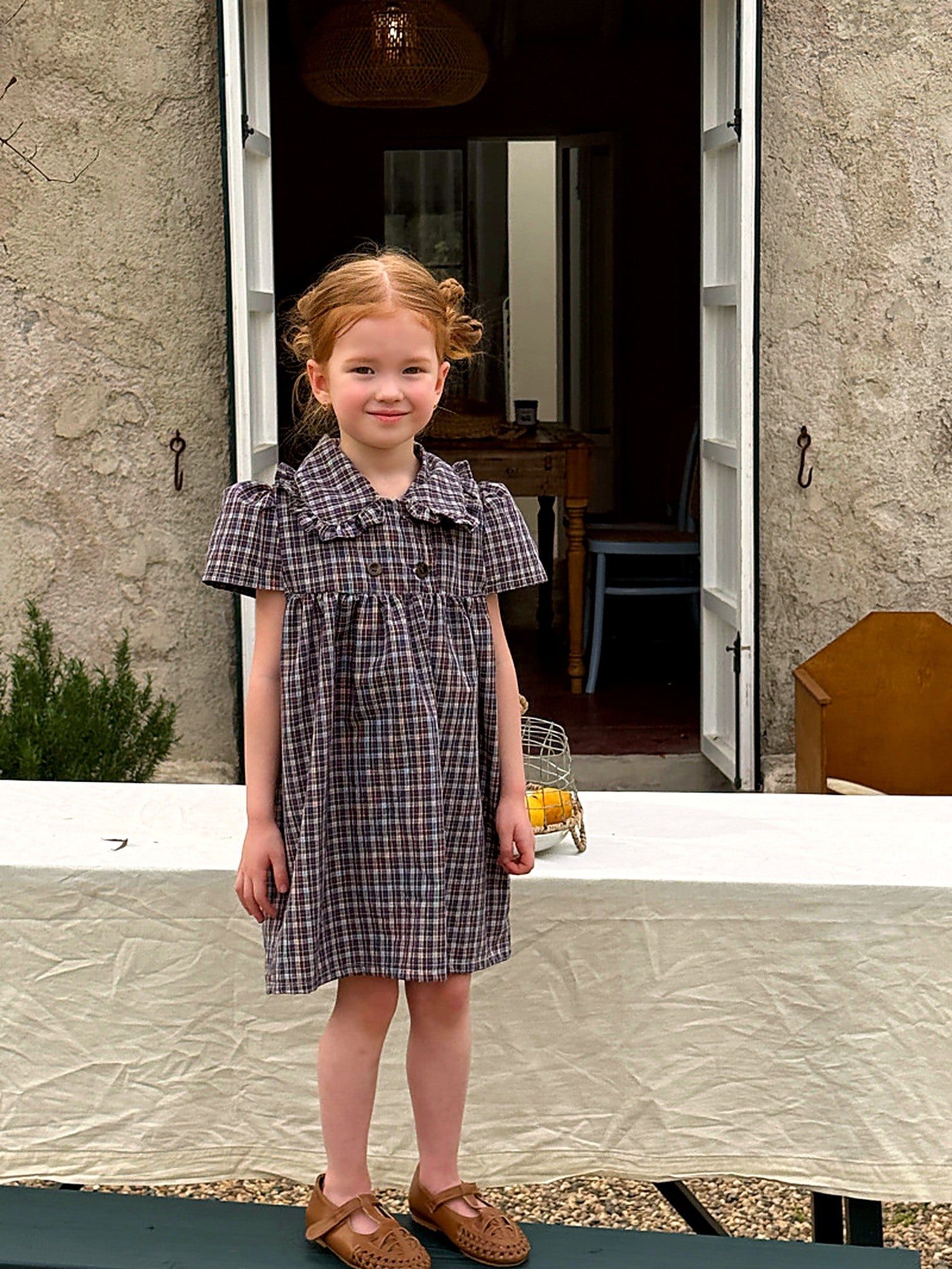 A girl in a blue gingham cotton dress with a doll collar, in a sunlit courtyard.