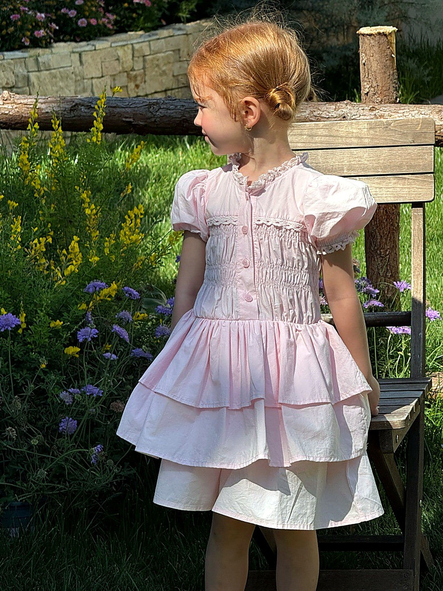 A girl in a pink cotton dress with a smocked bodice and puff sleeves, in a sunlit garden.