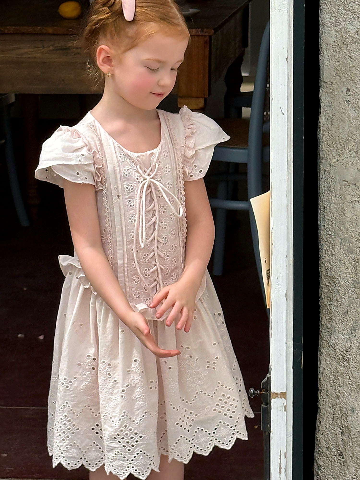 A girl in a faded-pink cotton eyelet dress with puff sleeves, by a stone wall in soft light.