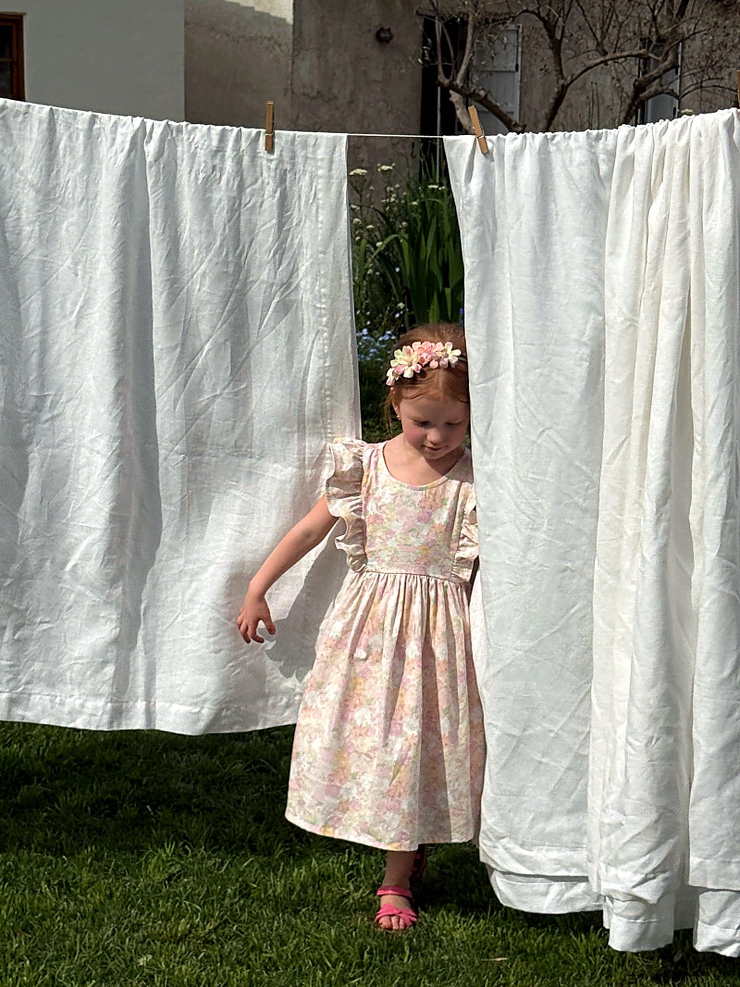 A girl in a blush floral cotton Peony Dress, walking past white linen hung to dry in a sunlit garden.