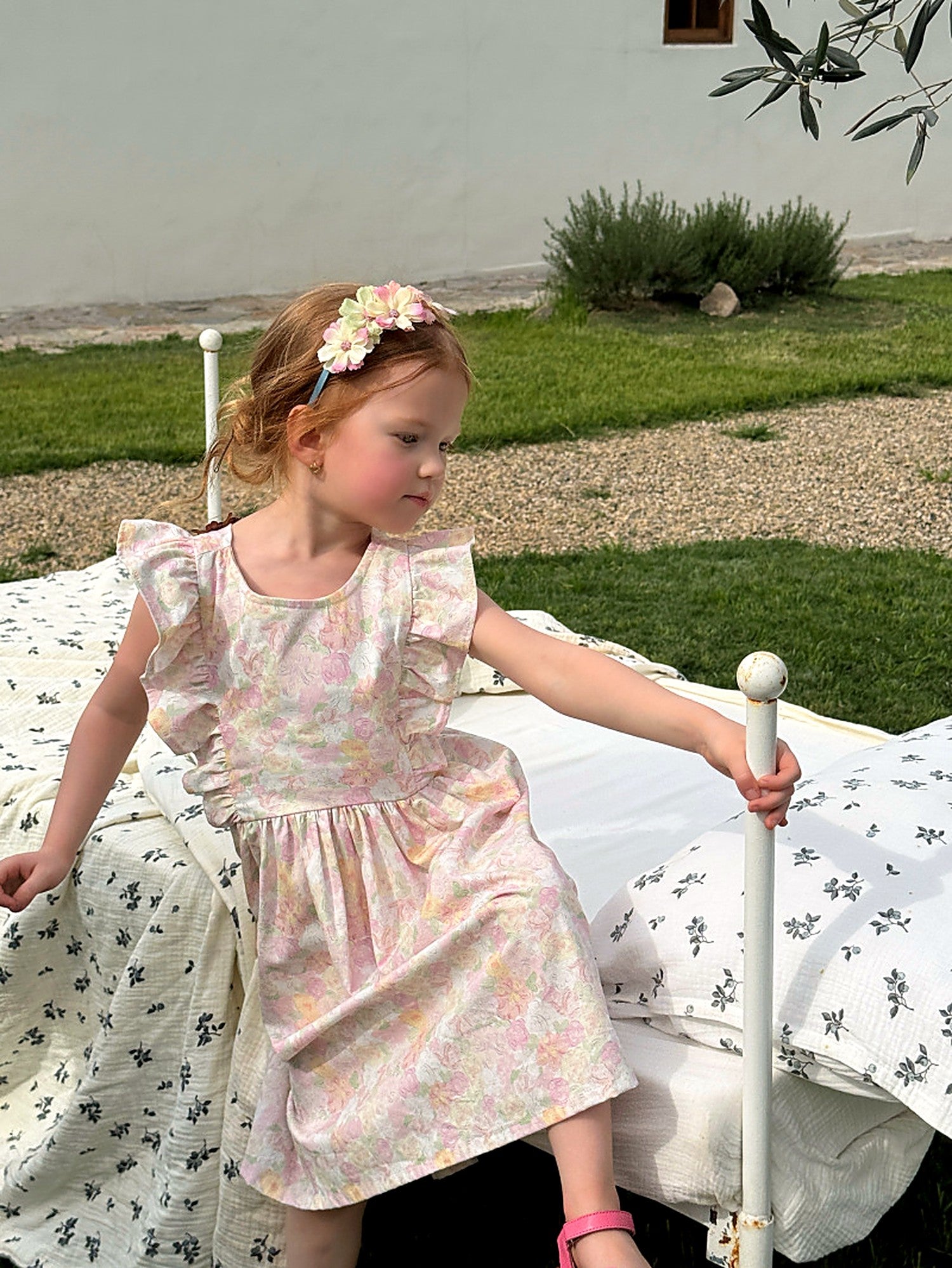 A girl in the Peony Dress resting on a white iron daybed set out in the grass.