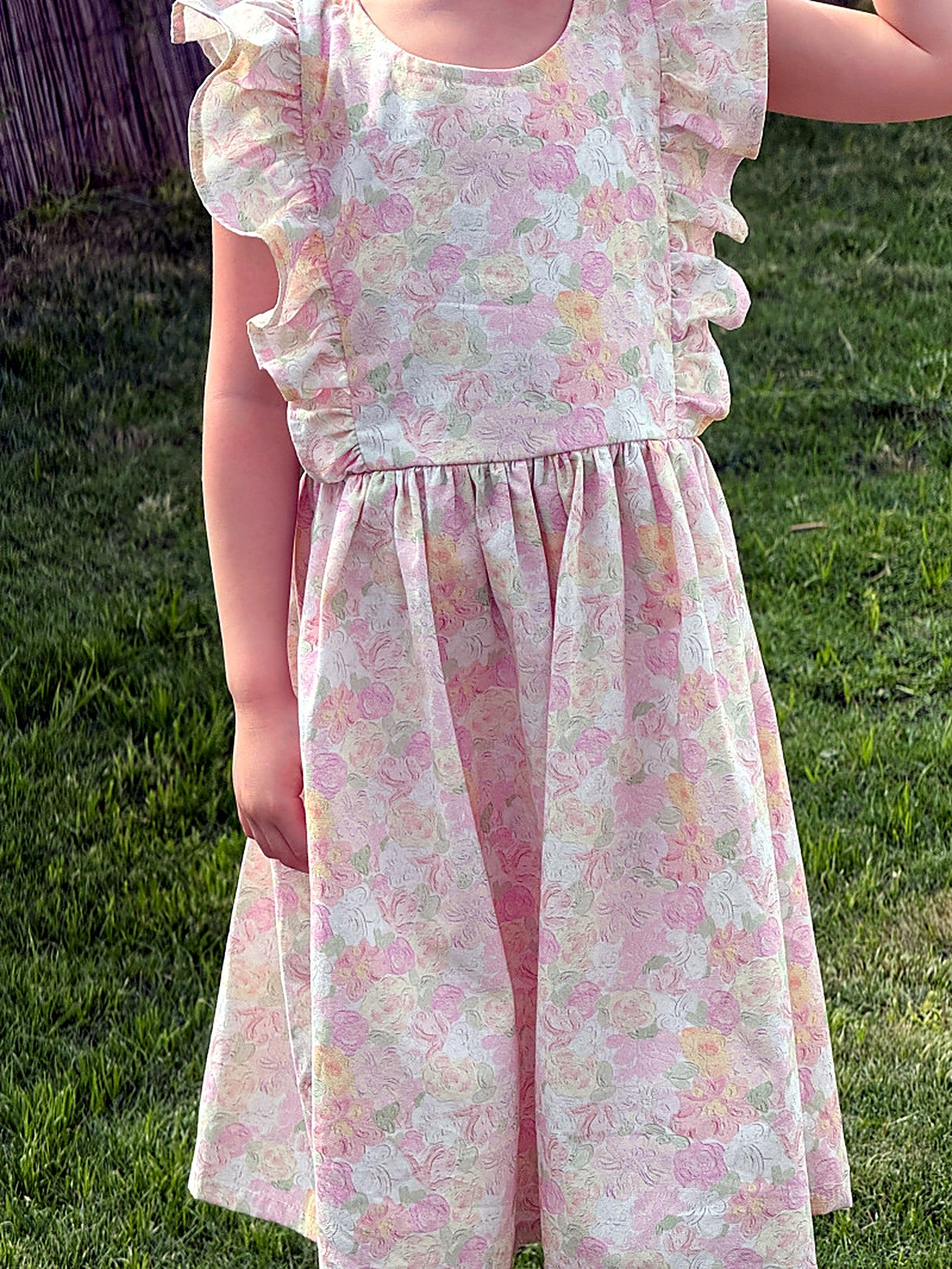 A child lying among soft linens in the blush floral Peony Dress on an unhurried afternoon.