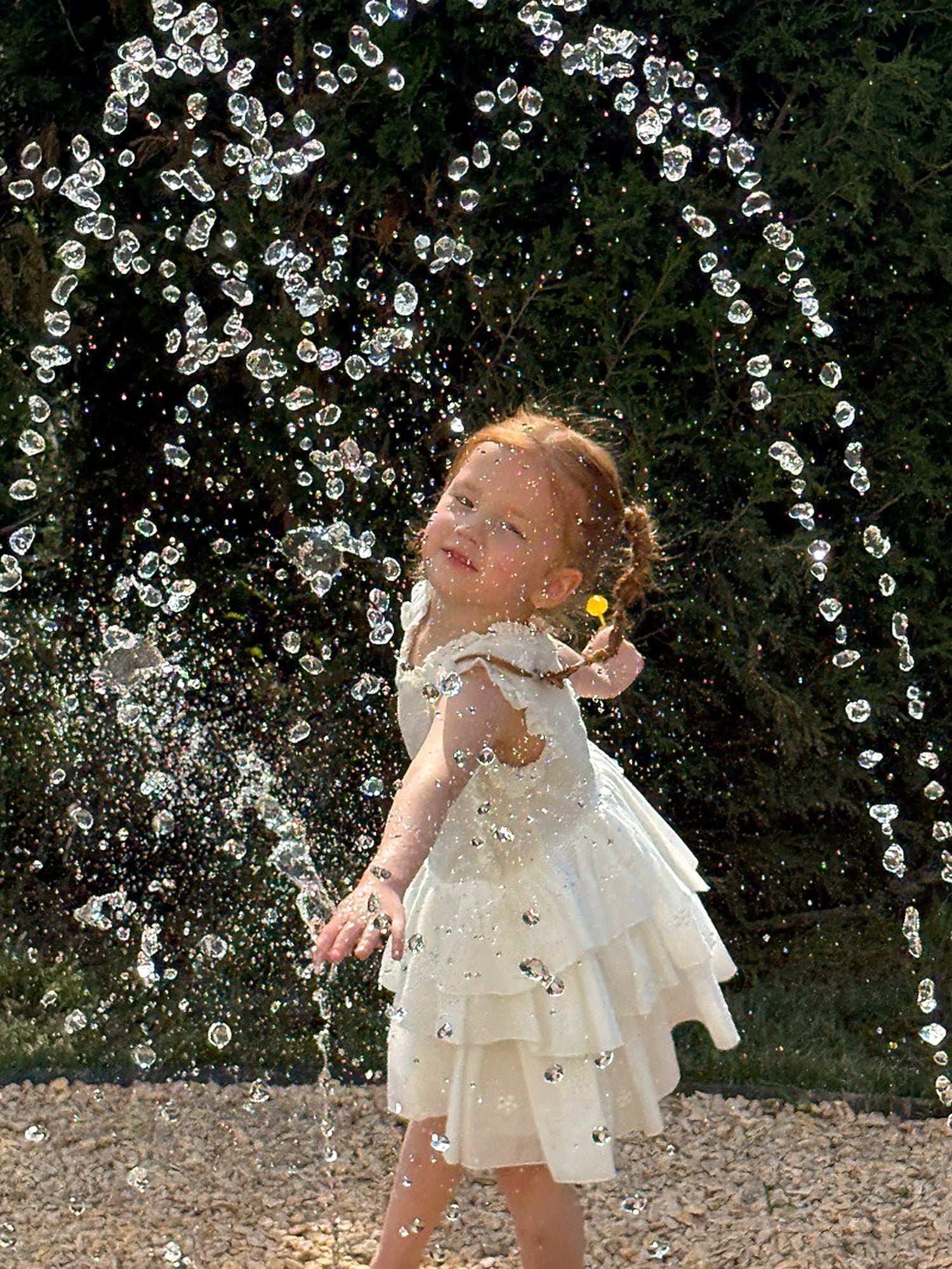 A girl in the Snowdrop Dress by a garden fountain, soft afternoon light.